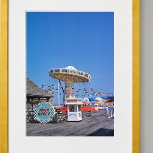May include: A vintage photograph of a carnival ride called "Down Under" with a swinging carousel, a ticket booth, and a wooden boardwalk. The sky is a bright blue.