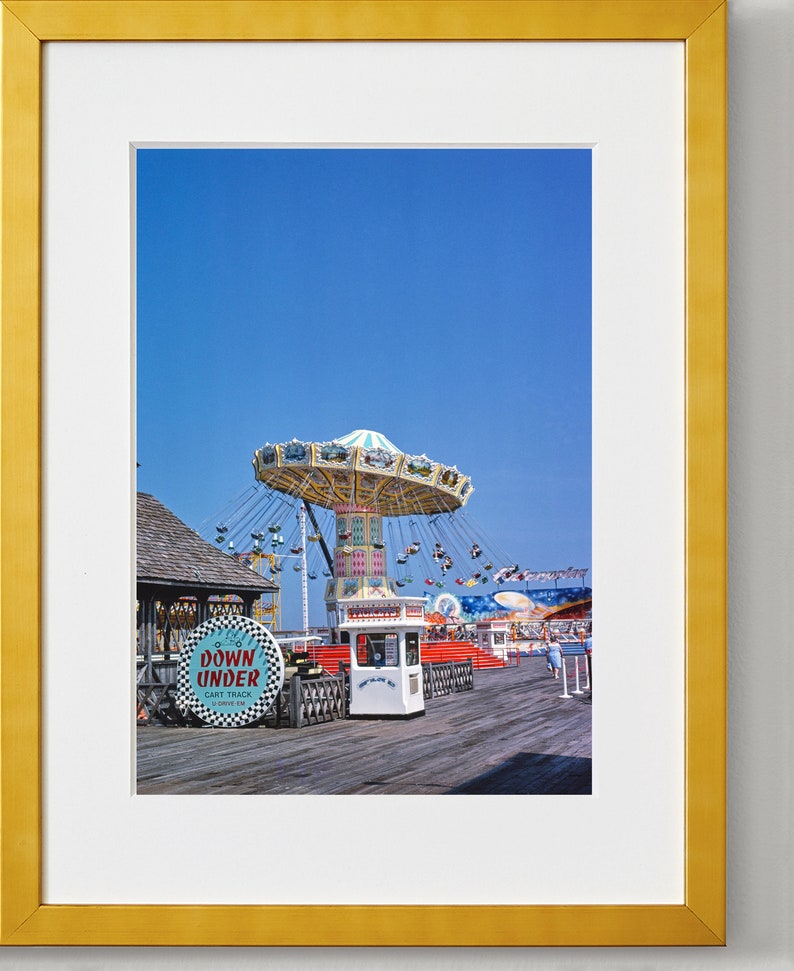 May include: A vintage photograph of a carnival ride called "Down Under" with a swinging carousel, a ticket booth, and a wooden boardwalk. The sky is a bright blue.