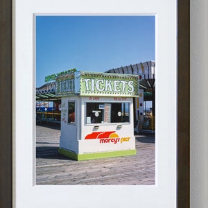 May include: A framed photograph of a ticket booth on a pier. The booth is white with green and red accents, and the sign reads "TICKETS." The booth has a window and the words "morcy's pier" are printed on the front. The sky is blue.