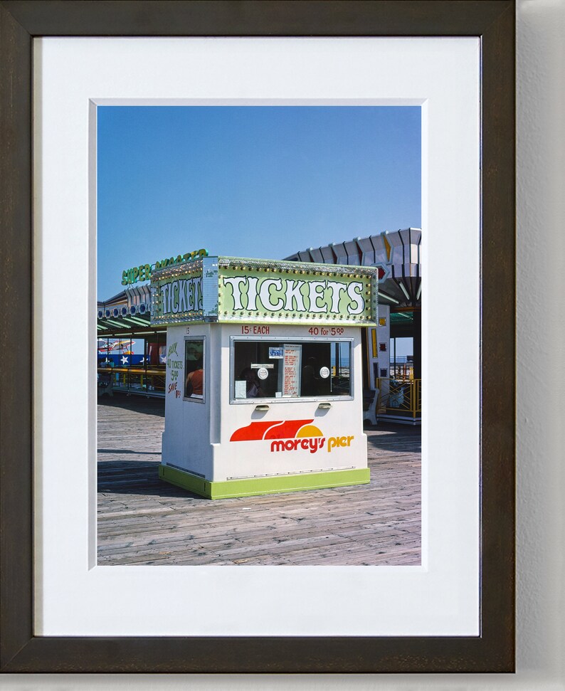 May include: A framed photograph of a ticket booth on a pier. The booth is white with green and red accents, and the sign reads "TICKETS." The booth has a window and the words "morcy's pier" are printed on the front. The sky is blue.