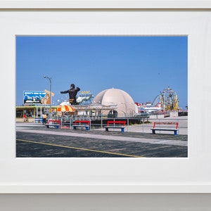 Op de afbeelding: Een vintage foto van een pretpark aan een boulevard met een groot gorillabeeld, een wit koepelvormig gebouw en een achtbaan op de achtergrond. Op de voorgrond staan banken en een bord met de tekst "Monkey's King of the Jungle".