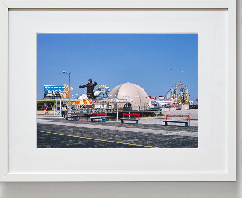 Op de afbeelding: Een vintage foto van een pretpark aan een boulevard met een groot gorillabeeld, een wit koepelvormig gebouw en een achtbaan op de achtergrond. Op de voorgrond staan banken en een bord met de tekst "Monkey's King of the Jungle".