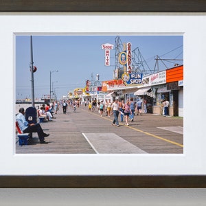 Peut inclure: Une photographie vintage d'une promenade avec des gens qui marchent et s'assoient sur des bancs. Il y a plusieurs magasins et restaurants avec des enseignes color&eacute;es, dont "Snow White", "Grandpa's" et "Fun Shop".