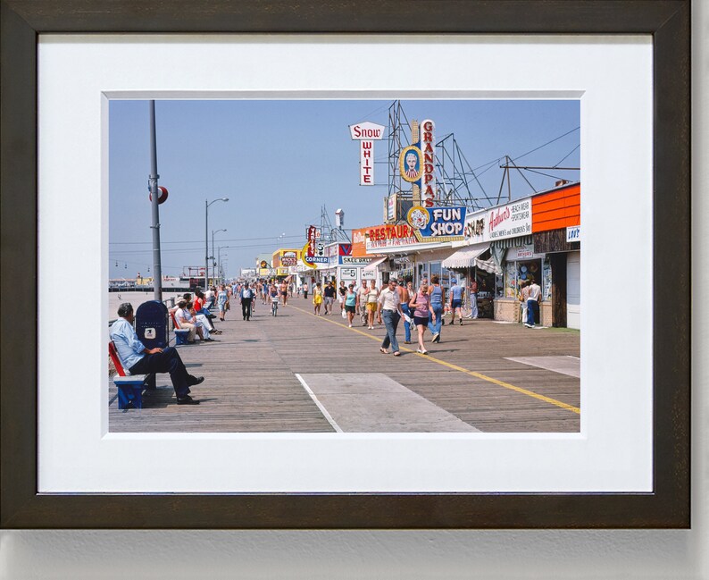 Peut inclure: Une photographie vintage d'une promenade avec des gens qui marchent et s'assoient sur des bancs. Il y a plusieurs magasins et restaurants avec des enseignes color&eacute;es, dont "Snow White", "Grandpa's" et "Fun Shop".