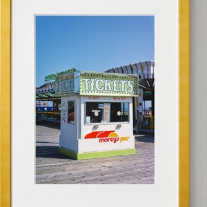 May include: A framed photograph of a ticket booth at an amusement park. The booth is white with green and red accents, and the sign reads "TICKETS." The booth is on a wooden pier under a clear blue sky.