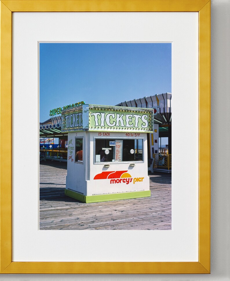 May include: A framed photograph of a ticket booth at an amusement park. The booth is white with green and red accents, and the sign reads "TICKETS." The booth is on a wooden pier under a clear blue sky.