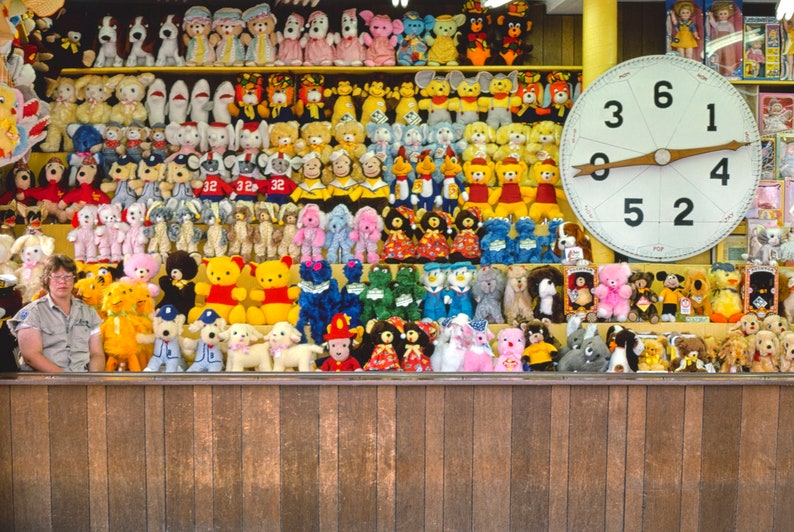 May include: A carnival game with a large spinning wheel and a wall of stuffed animals. The wheel has numbers 0, 1, 2, 3, 4, 5, and 6. The stuffed animals are mostly in shades of pink, blue, yellow, and green.