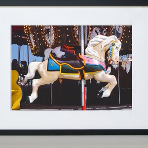 May include: A white carousel horse with a colorful saddle and a blue and gold bridle. The horse is in motion, with its legs raised in the air. The carousel is decorated with lights and other horses.