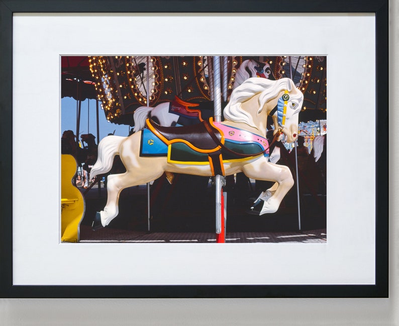 May include: A white carousel horse with a colorful saddle and a blue and gold bridle. The horse is in motion, with its legs raised in the air. The carousel is decorated with lights and other horses.