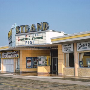 K&ouml;nnte beinhalten: Ein gelbes und wei&szlig;es Geb&auml;ude mit einem Schild, auf dem "STRAND" und "SYLVESTER STALLONE FIST" in roten und blauen Buchstaben steht. Das Geb&auml;ude hat ein gro&szlig;es Fenster mit einem Schild, auf dem "Spiro's RESTAURANT" in schwarzen Buchstaben steht.