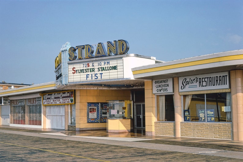 K&ouml;nnte beinhalten: Ein gelbes und wei&szlig;es Geb&auml;ude mit einem Schild, auf dem "STRAND" und "SYLVESTER STALLONE FIST" in roten und blauen Buchstaben steht. Das Geb&auml;ude hat ein gro&szlig;es Fenster mit einem Schild, auf dem "Spiro's RESTAURANT" in schwarzen Buchstaben steht.