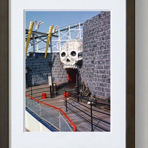 Puede incluir: Una escultura de calavera en blanco y negro con una boca roja, sobre un fondo de pared de ladrillo gris. La calavera es parte de una atracci&oacute;n de parque de atracciones, con una pasarela de madera y barandillas de metal negro que conducen a la entrada.