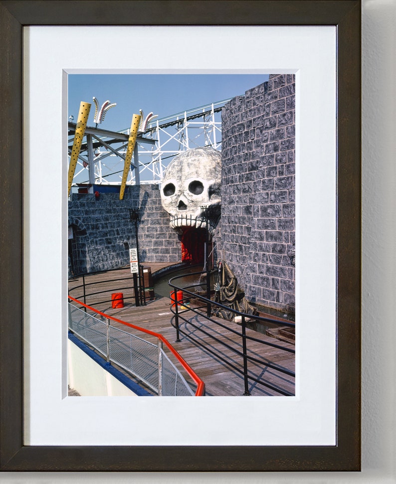 Puede incluir: Una escultura de calavera en blanco y negro con una boca roja, sobre un fondo de pared de ladrillo gris. La calavera es parte de una atracci&oacute;n de parque de atracciones, con una pasarela de madera y barandillas de metal negro que conducen a la entrada.