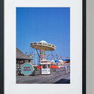 May include: A vintage photograph of a fairground ride on a pier. The ride is a carousel with a white and blue canopy and colourful seats. The ride is surrounded by other fairground attractions, including a small building with a sign that reads "Down Under Cart Track". The sky is blue and the pier is made of wood.
