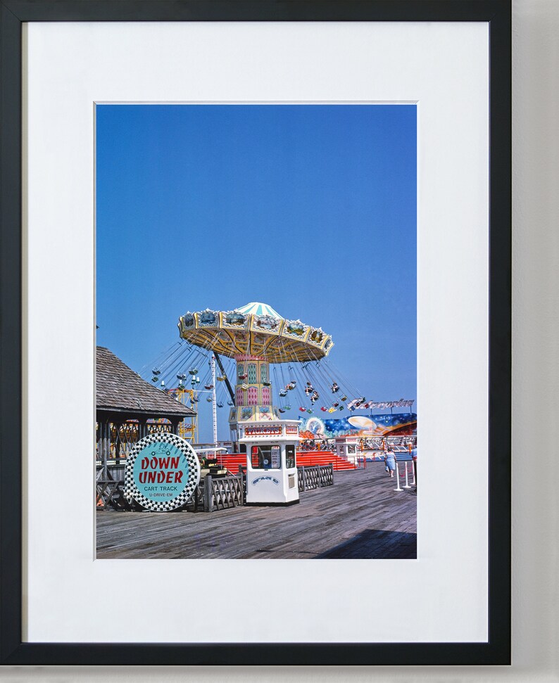 May include: A vintage photograph of a fairground ride on a pier. The ride is a carousel with a white and blue canopy and colourful seats. The ride is surrounded by other fairground attractions, including a small building with a sign that reads "Down Under Cart Track". The sky is blue and the pier is made of wood.