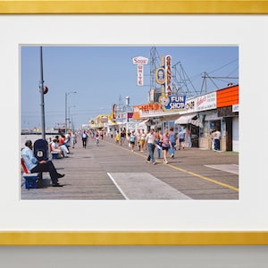 Peut inclure: Une photographie vintage d'une promenade avec des gens qui marchent et s'assoient sur des bancs. Il y a plusieurs magasins et restaurants avec des enseignes color&eacute;es, dont "Snow White", "Grandpa's", "Fun Shop" et "Restaurant".