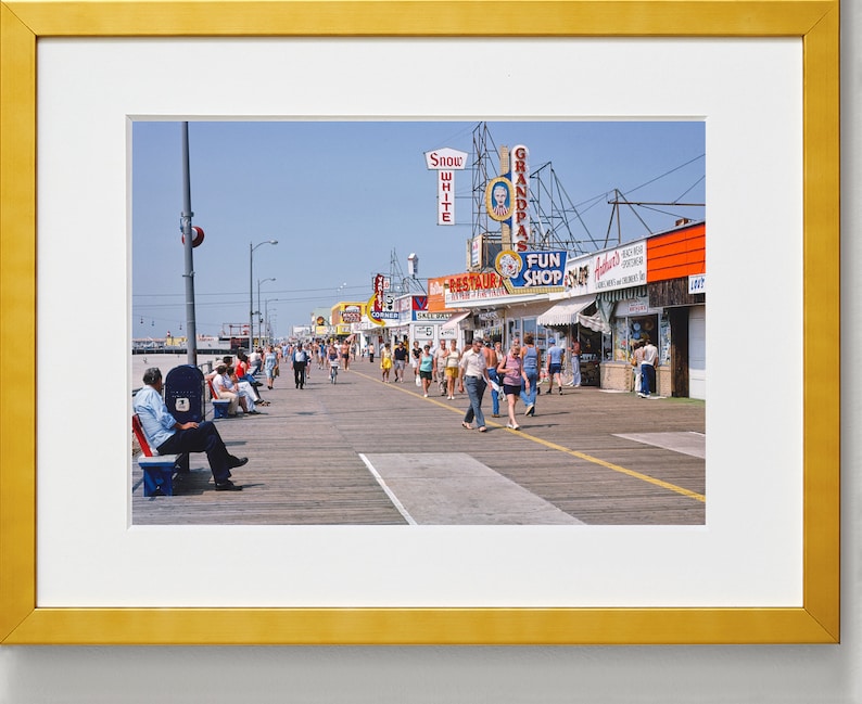 Peut inclure: Une photographie vintage d'une promenade avec des gens qui marchent et s'assoient sur des bancs. Il y a plusieurs magasins et restaurants avec des enseignes color&eacute;es, dont "Snow White", "Grandpa's", "Fun Shop" et "Restaurant".