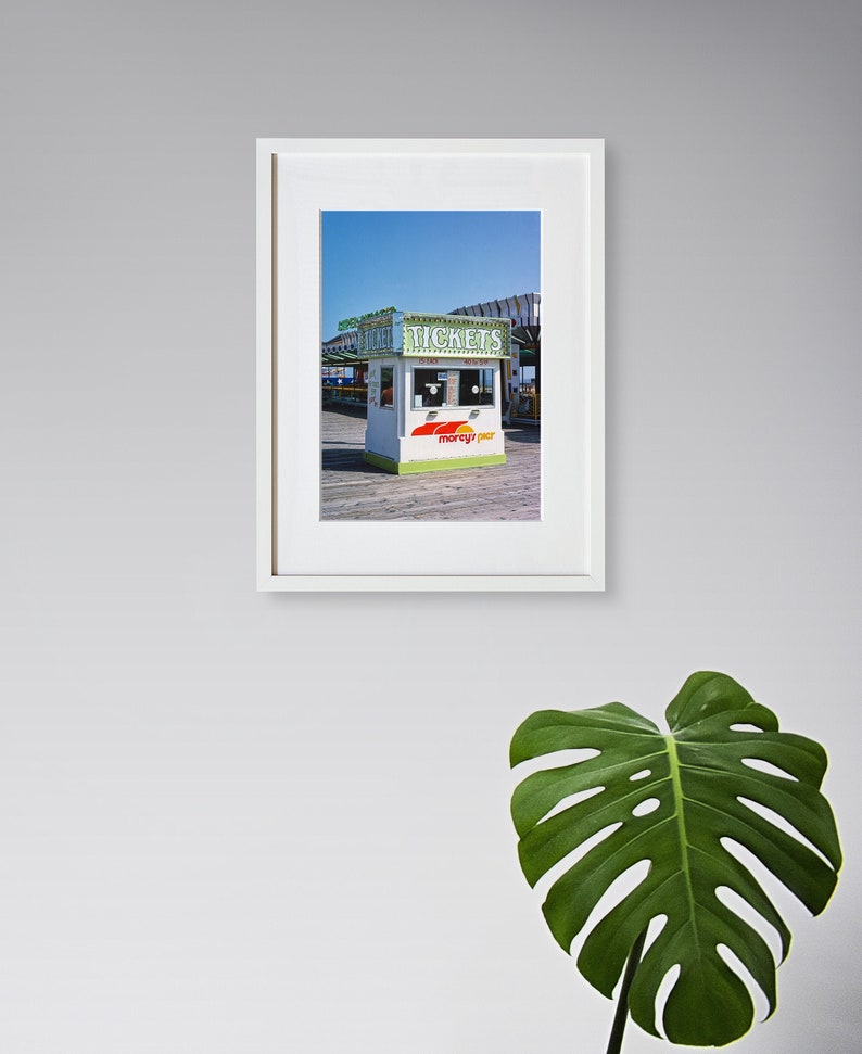 May include: Framed print of a ticket booth on a pier, with the word "TICKETS" visible. The booth is white with green and red accents, set against a blue sky. The print is in a white frame.