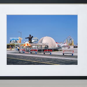 Op de afbeelding: Een vintage foto van een pretpark aan een boulevard met een groot gorillabeeld, een wit koepelvormig gebouw en een achtbaan op de achtergrond. Er zijn banken en een bord met de tekst "Monkeys King of the Jungle".