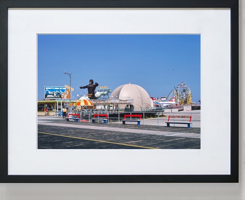 Op de afbeelding: Een vintage foto van een pretpark aan een boulevard met een groot gorillabeeld, een wit koepelvormig gebouw en een achtbaan op de achtergrond. Er zijn banken en een bord met de tekst "Monkeys King of the Jungle".