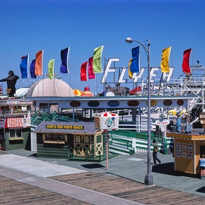 Puede incluir: Una escena de parque de atracciones vintage con una monta&ntilde;a rusa, un gran letrero que dice "Flyer" y varias banderas de colores. Hay varias tiendas peque&ntilde;as y quioscos a lo largo de un paseo mar&iacute;timo, con gente caminando.