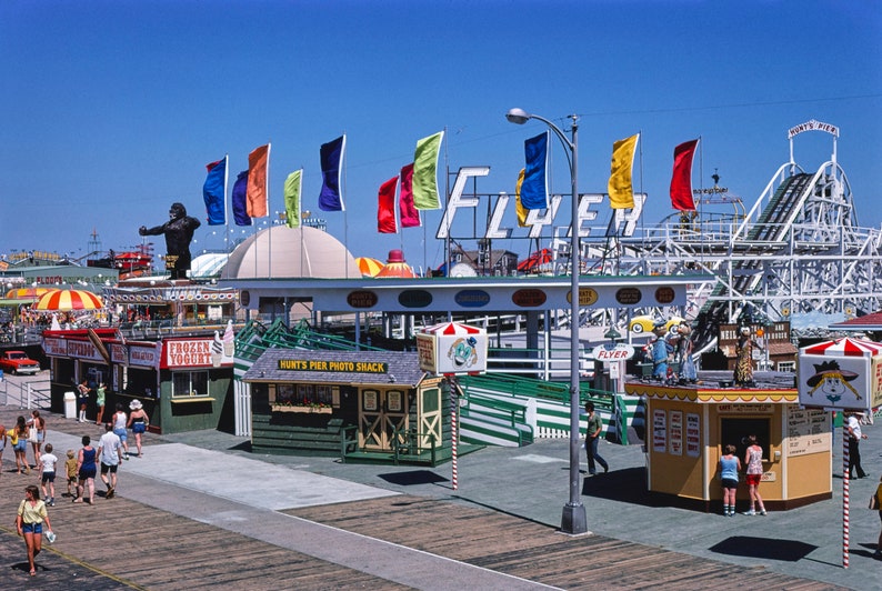 Puede incluir: Una escena de parque de atracciones vintage con una monta&ntilde;a rusa, un gran letrero que dice "Flyer" y varias banderas de colores. Hay varias tiendas peque&ntilde;as y quioscos a lo largo de un paseo mar&iacute;timo, con gente caminando.