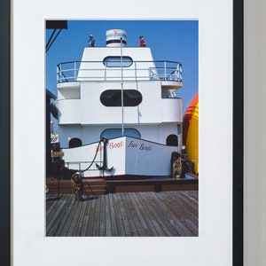 May include: A white boat with a red and blue sign that says "Fun Boat" is docked at a pier. The boat has a small upper deck with a railing and a red flag on top.