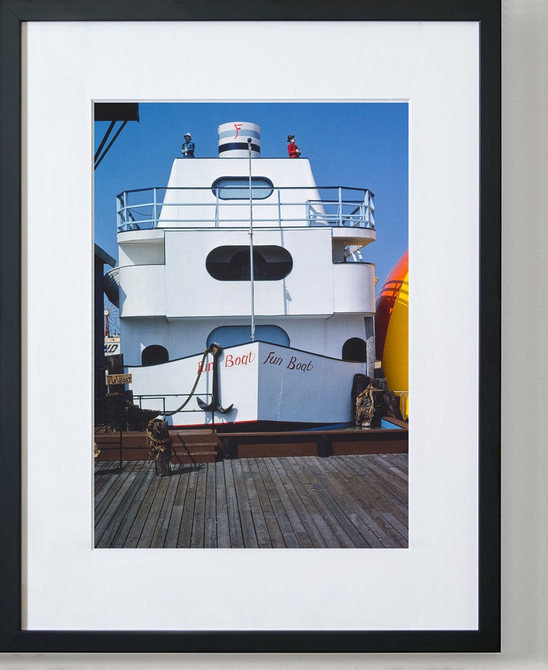 May include: A white boat with a red and blue sign that says "Fun Boat" is docked at a pier. The boat has a small upper deck with a railing and a red flag on top.