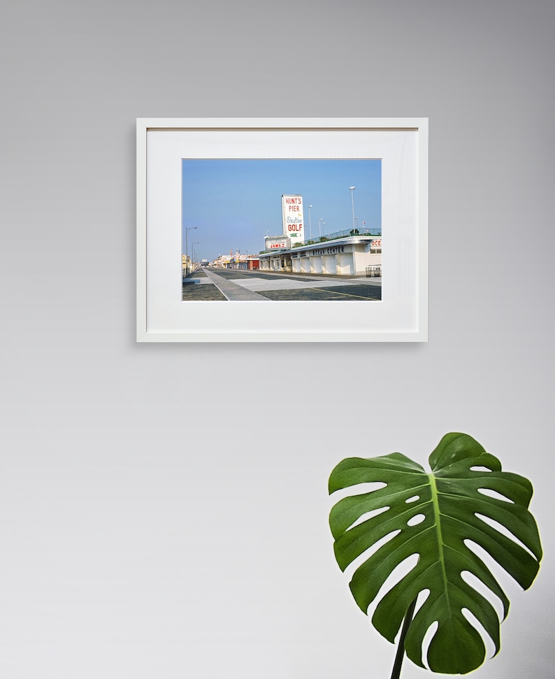 May include: A vintage photograph of a building with a sign that reads "Hunt's Pier Putting Golf" in red letters. The building is located on a boardwalk with a blue sky in the background.