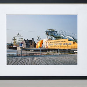 May include: A vintage photograph of a boardwalk amusement park with a yellow sign that reads "Morey's Pier Presents... Bear Country Jubilee". The sign is on a building with a large, colorful, inflatable bear. The Wipeout ride is visible in the background.