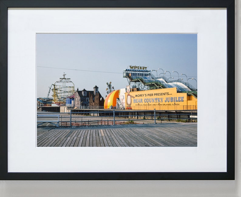May include: A vintage photograph of a boardwalk amusement park with a yellow sign that reads "Morey's Pier Presents... Bear Country Jubilee". The sign is on a building with a large, colorful, inflatable bear. The Wipeout ride is visible in the background.