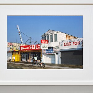 May include: A brightly colored storefront with a red and yellow sign that reads "Skilo" and a red sign that reads "Nates's Frankfurters". The storefront is located on a boardwalk with a blue sky in the background. There are people riding bicycles in front of the store.
