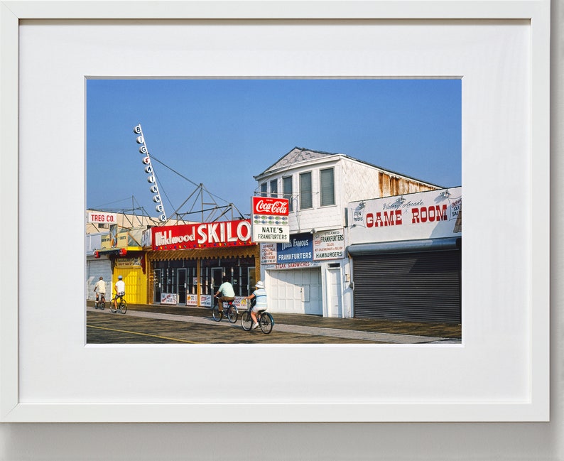 May include: A brightly colored storefront with a red and yellow sign that reads "Skilo" and a red sign that reads "Nates's Frankfurters". The storefront is located on a boardwalk with a blue sky in the background. There are people riding bicycles in front of the store.