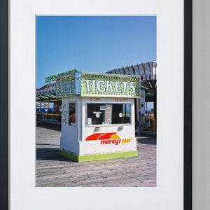 May include: A white ticket booth with green and red accents, labeled "TICKETS" in large letters, stands on a wooden pier. The booth has a sign that says "morcy's pier" and is set against a clear blue sky. A carousel is visible in the background.
