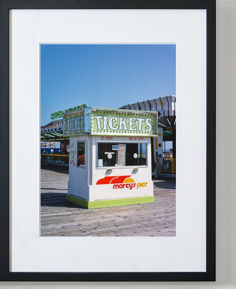 May include: A white ticket booth with green and red accents, labeled "TICKETS" in large letters, stands on a wooden pier. The booth has a sign that says "morcy's pier" and is set against a clear blue sky. A carousel is visible in the background.