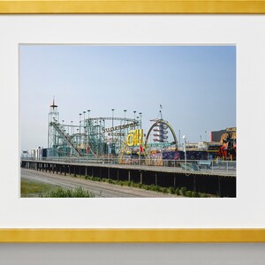 Op de afbeelding: Een ingelijste foto van een pretpark met een achtbaan en andere attracties. De achtbaan is groen en zilverkleurig, met de woorden "Supersonic Roller Coaster" zichtbaar. De lucht is helderblauw.