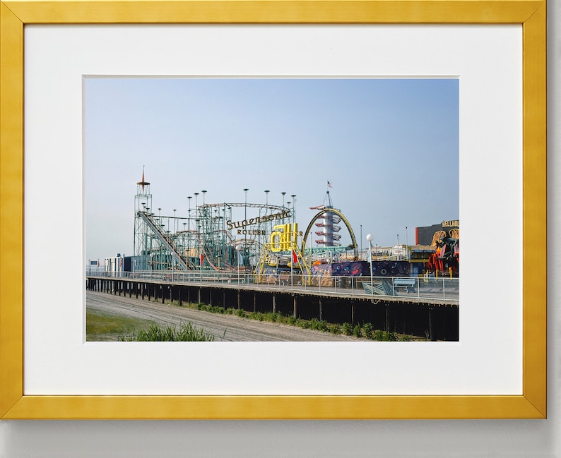 Op de afbeelding: Een ingelijste foto van een pretpark met een achtbaan en andere attracties. De achtbaan is groen en zilverkleurig, met de woorden "Supersonic Roller Coaster" zichtbaar. De lucht is helderblauw.