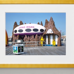 May include: A white and purple spaceship with the text "STAR WARS" on the side, surrounded by brown rock formations. Two yellow figures stand in front of the spaceship, one resembling a robot and the other a human.