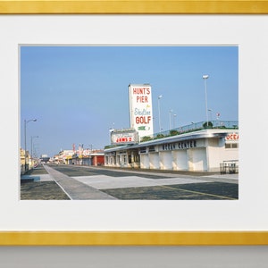 May include: A vintage photograph of a street scene in a seaside town. The photo shows a street lined with shops and businesses, including a shop called "Jaws 2" and a building with a sign that reads "Hunt's Pier Skyline Golf".