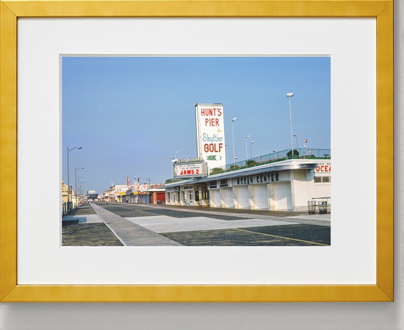 May include: A vintage photograph of a street scene in a seaside town. The photo shows a street lined with shops and businesses, including a shop called "Jaws 2" and a building with a sign that reads "Hunt's Pier Skyline Golf".