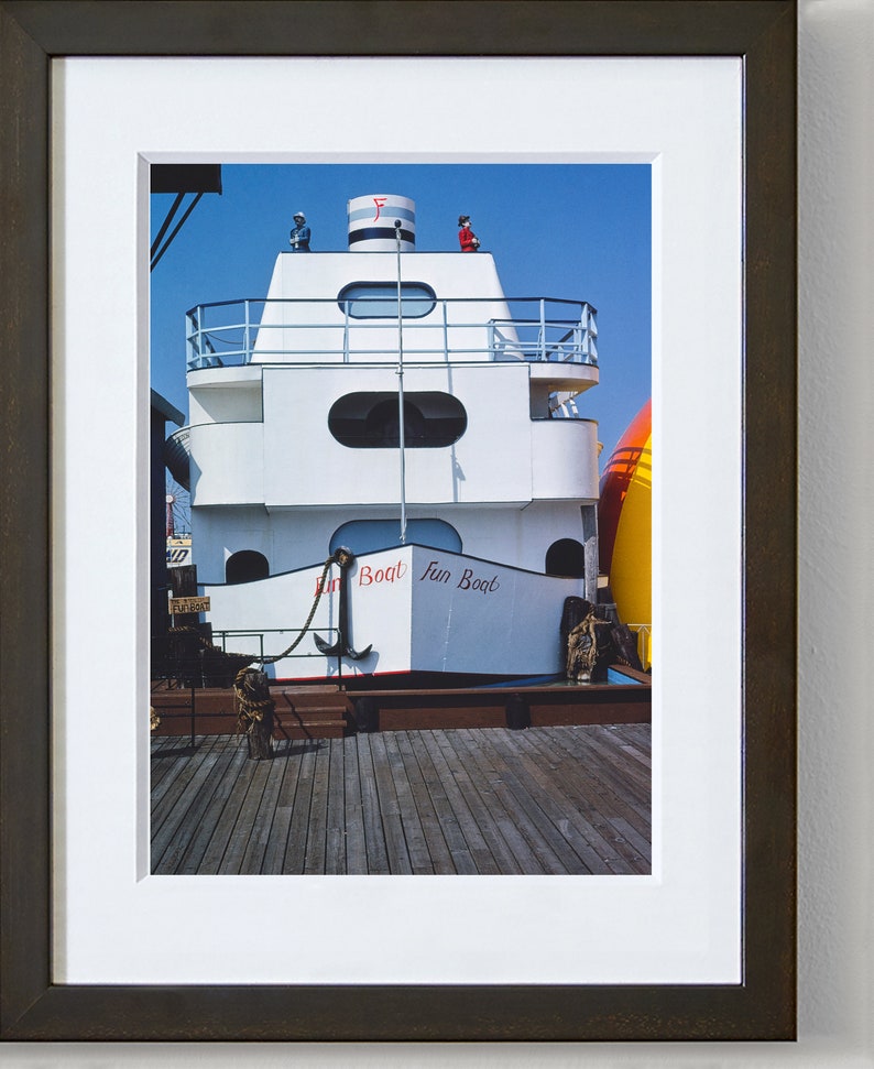 May include: A white boat with red and blue accents, docked at a pier. The boat has a large anchor hanging from the front and the words "Fun Boat" written on the side.