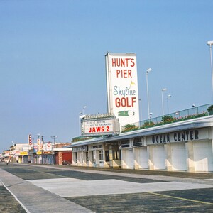 May include: A vintage photograph of a boardwalk with a movie theatre marquee advertising "Jaws 2" and a sign for "Hunt's Pier Skyline Golf".