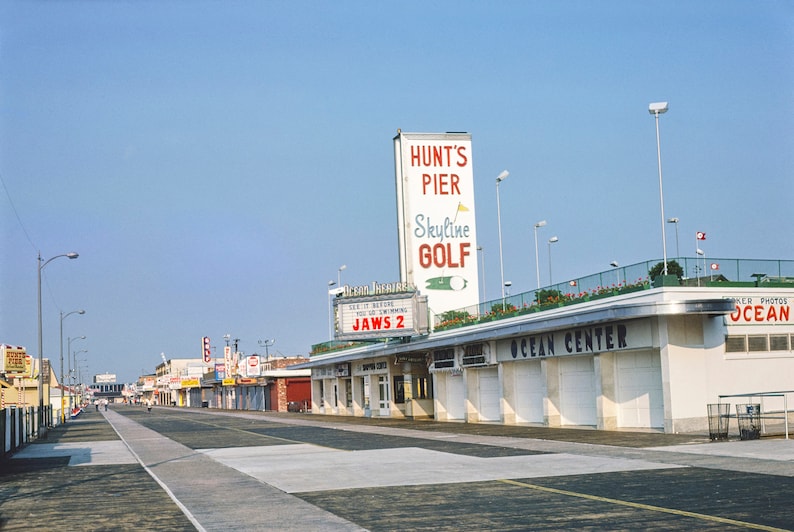 May include: A vintage photograph of a boardwalk with a movie theatre marquee advertising "Jaws 2" and a sign for "Hunt's Pier Skyline Golf".