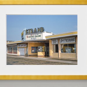 K&ouml;nnte beinhalten: Ein Vintage-Foto eines Kinos namens "The Strand" mit einem Leuchtkasten, der den Film "Fist" mit Sylvester Stallone bewirbt. Neben dem Kino befindet sich ein Restaurant namens "Spire's Restaurant".