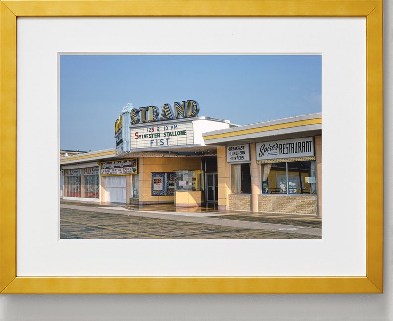 K&ouml;nnte beinhalten: Ein Vintage-Foto eines Kinos namens "The Strand" mit einem Leuchtkasten, der den Film "Fist" mit Sylvester Stallone bewirbt. Neben dem Kino befindet sich ein Restaurant namens "Spire's Restaurant".