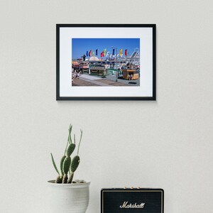 Puede incluir: Una fotograf&iacute;a enmarcada de un parque de atracciones vintage con banderas de colores y una monta&ntilde;a rusa en el fondo. La foto est&aacute; colocada contra una pared blanca con una planta de cactus y un altavoz negro Marshall en primer plano.
