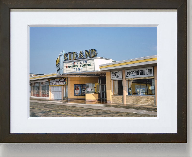 K&ouml;nnte beinhalten: Ein Vintage-Foto eines Kinos namens "The Strand" mit einem Leuchtkasten, der "Sylvester Stallone Fist" um 19:25 und 22:00 Uhr bewirbt.  Nebenan befindet sich ein Restaurant namens "Spire's Restaurant".