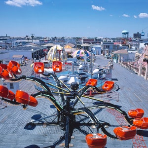 May include: A carnival ride with orange seats on a black metal frame. The ride is on a wooden boardwalk with other carnival rides and buildings in the background.