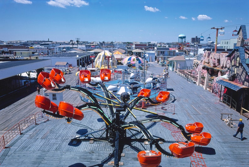 May include: A carnival ride with orange seats on a black metal frame. The ride is on a wooden boardwalk with other carnival rides and buildings in the background.