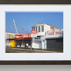 May include: A vintage photograph of a boardwalk scene with a red and yellow sign that reads "Wildwood Skillo". There are four people riding bicycles in front of the sign. The sign is in front of a building with a Coca-Cola sign and a sign that reads "Game Room".